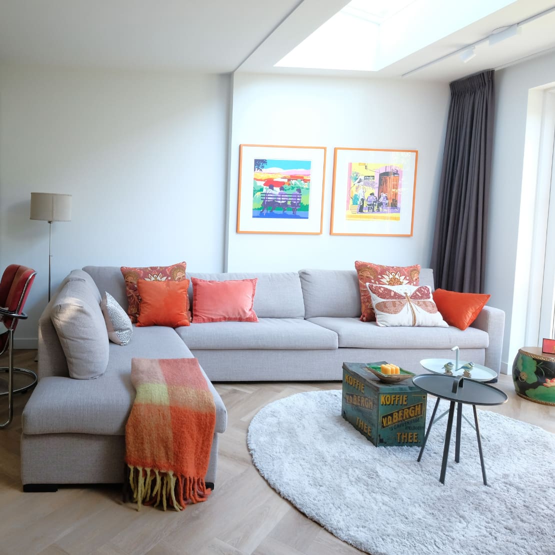 Interieurstyling woonkamer met kleurrijke accessoires en kunst aan de wand door Kirsten Hermsen Gestylde woonkamer met grijze hoekbank, oranje kussens en kleurrijke kunst aan de wand